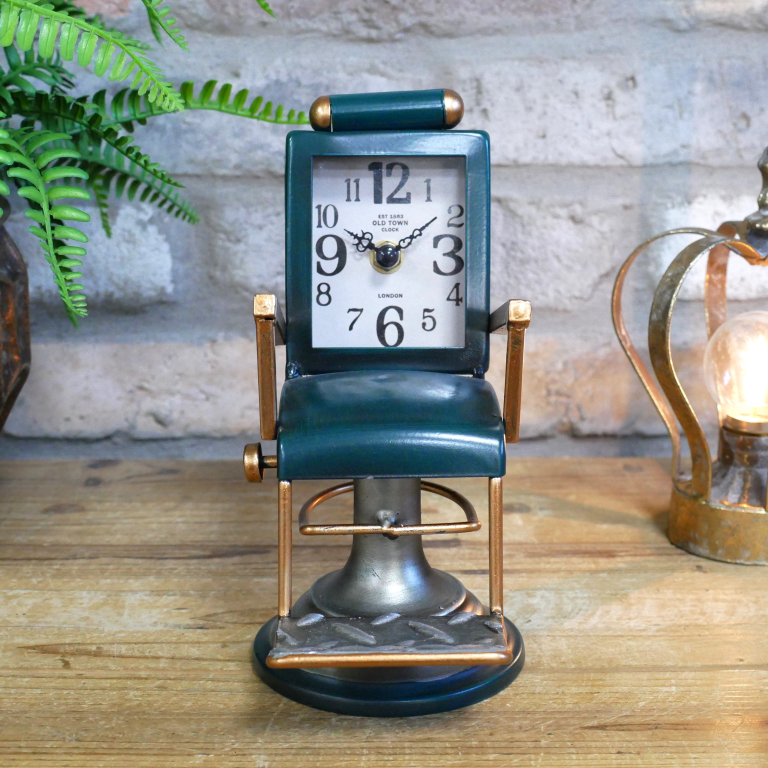 Vintage-style clock in the shape of a barber chair on a wooden surface with a plant and lamp in the background.
