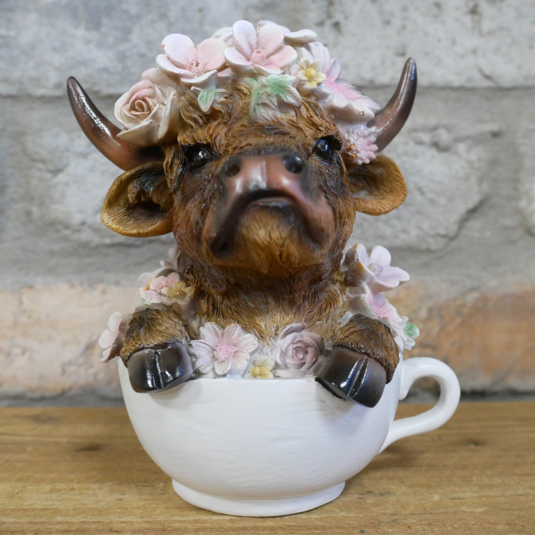 Decorative figurine of a highland cow with flowers sitting inside a teacup on a wooden surface.