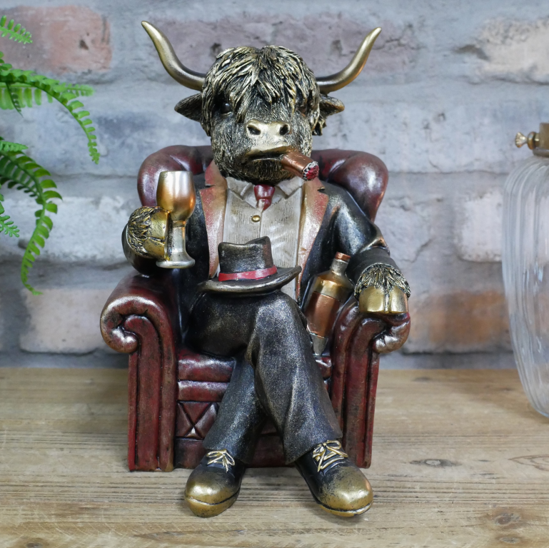 Statue of a highland cow in formal attire sitting in a chair with a glass and bottle, against a brick wall.